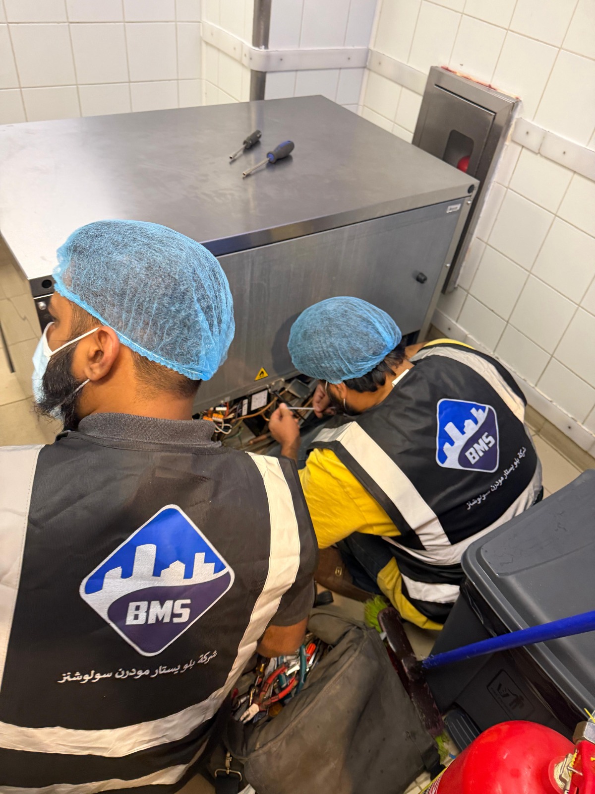 Two BMS technicians working on the exposed compressor compartment of an under-counter refrigerator