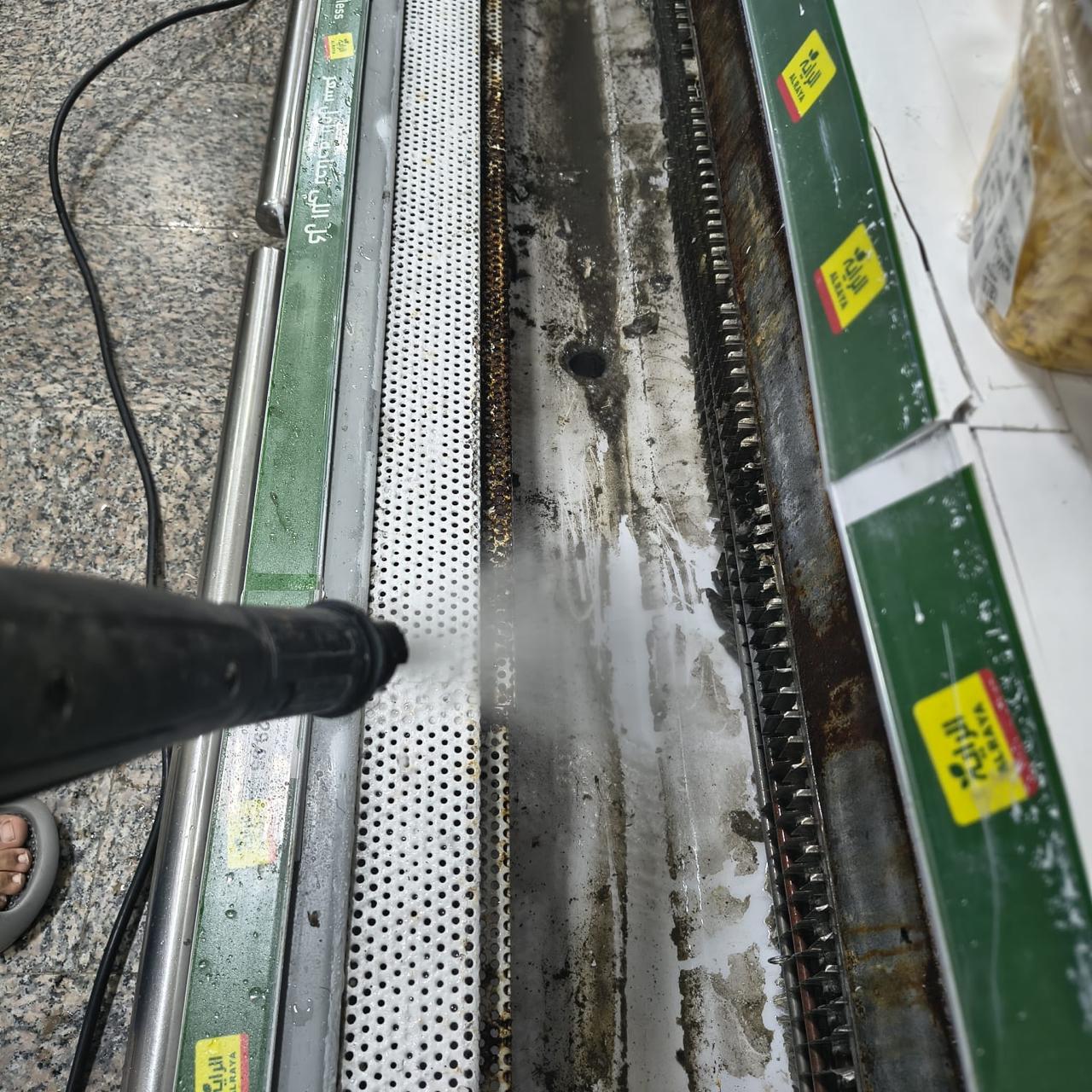 Pressure-washer lance spraying the base tray and evaporator fins of an open multideck display chiller